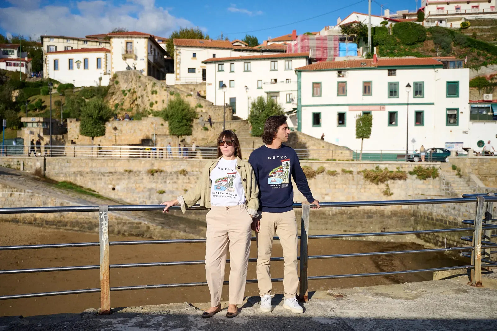 Una pareja con la sudadera Getxo · Puerto Viejo y una camiseta blanca, apoyados en la barandilla del Puerto Viejo de Getxo, con las casas del barrio detrás