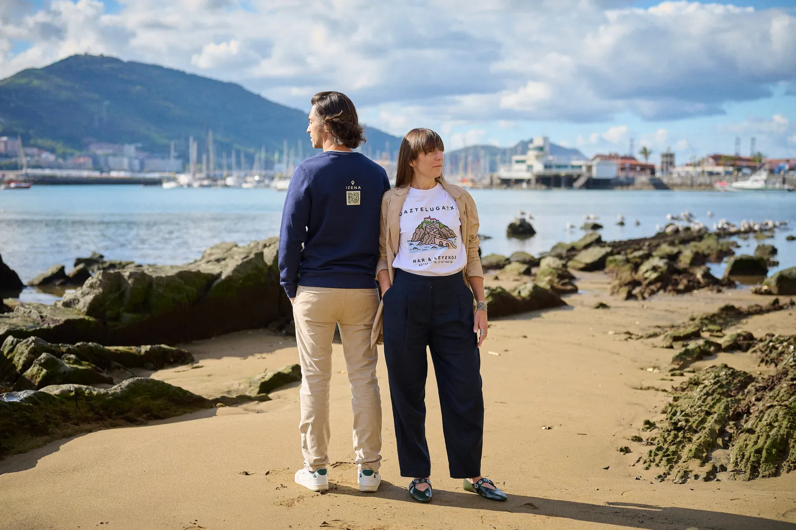 Una pareja con la sudadera azul marino y la camiseta Gaztelugatxe, de pie en la playa de Getxo, mirando al puerto