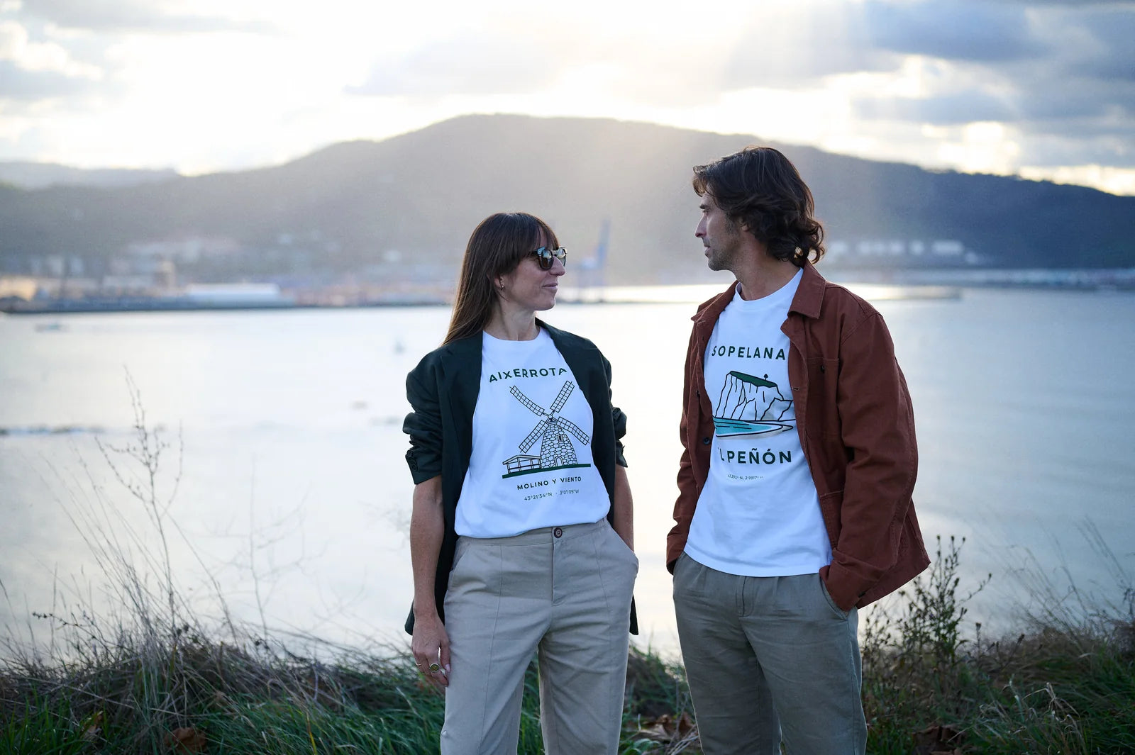 Una pareja con las camisetas blancas Aixerrota · Molino y Viento y Sopelana · El Peñón, mirándose junto a la ría de Getxo al atardecer
